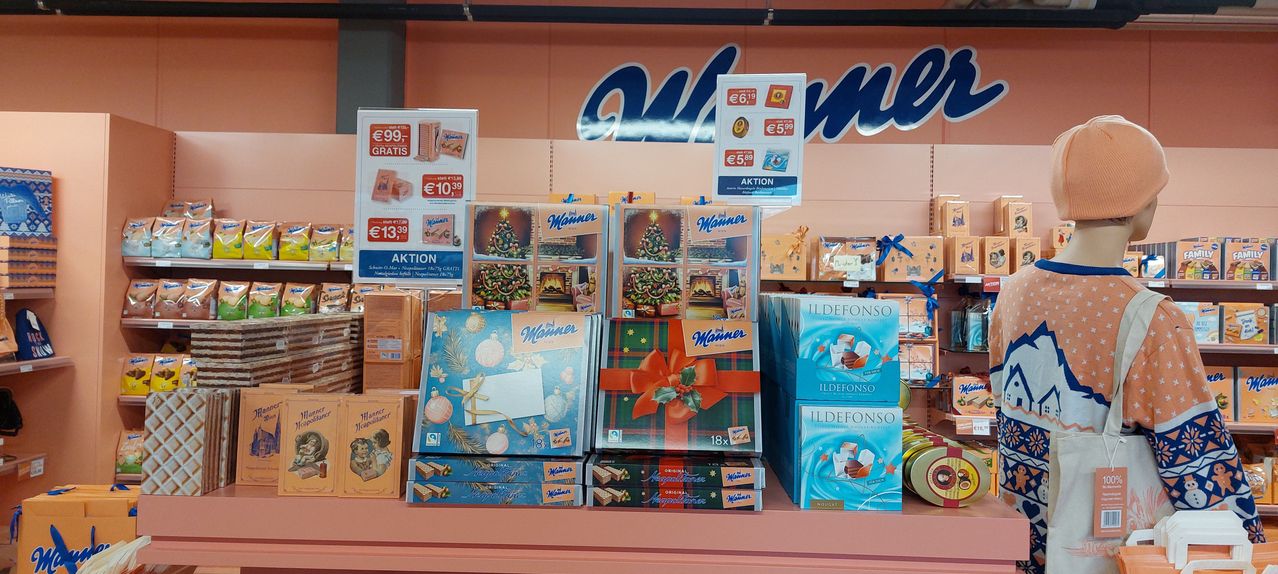 Eine Ladentheke mit verschiedenen Schachteln Schokoladen und anderen Produkten. Der Hintergrund zeigt ein blaues Schild mit großen weißen Buchstaben. Die Produkte umfassen Schachteln von 'Wanner' und 'Ildefonso'.