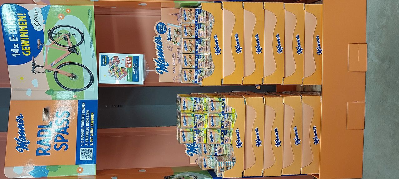Stapeln von gelben Schachteln mit der Aufschrift Manner Original und XXL-Pack sind ausgestellt. Im Vordergrund befindet sich ein Werbeschild mit einem Preis von 4,99 Euro.