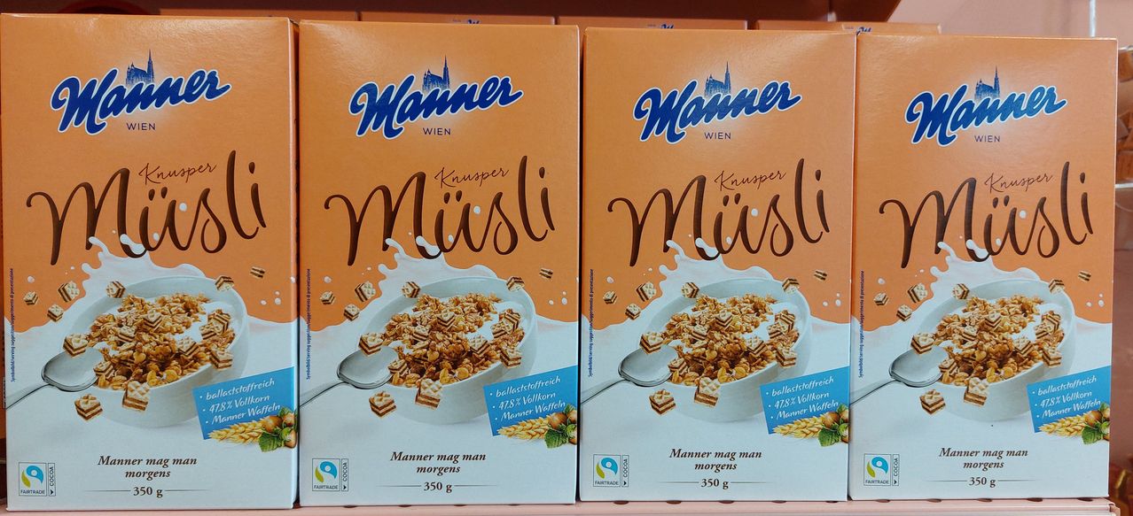 Zwei Schachteln Manner Muesli-Müsli, jede mit einer Schüssel Müsli und Milch, auf einem Regal ausgestellt. Das Müsli ist eine Mischung aus Waffel und Getreide mit einem blauen Etikett.