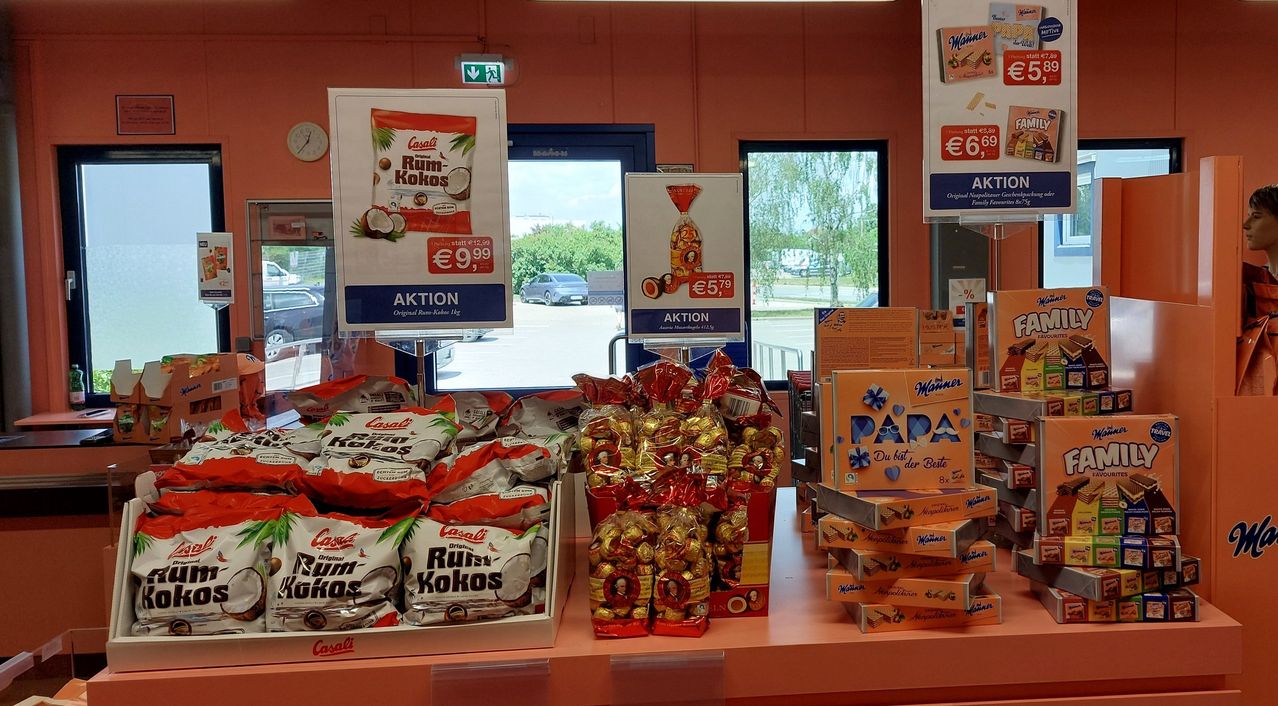 Store display with products like Rum-Kokos snacks and assorted chocolates on sale, with a clock and a car visible through the window.