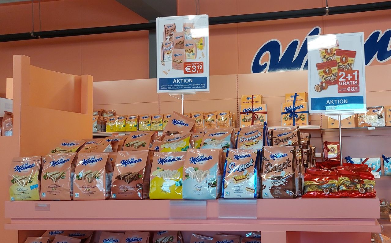 Ein Ladenregal zeigt zahlreiche Tüten mit Manner-Oblatenkeksen, einige in Schokolade und andere in Haselnussgeschmack. Ein Schild darüber bewirbt eine Aktion mit einem Preis von 3,19 Euro.