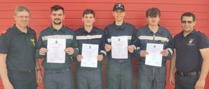 Fünf Männer in Uniform stehen vor einer roten Wand und halten Urkunden in den Händen. Der Mann auf der linken Seite trägt eine Uhr und einen blauen Gürtel. Die anderen Männer tragen schwarze Mützen.