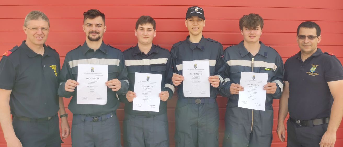 Fünf Männer in Uniform stehen vor einer roten Wand und halten Urkunden in den Händen. Der Mann auf der linken Seite trägt eine Uhr und einen blauen Gürtel. Die anderen Männer tragen schwarze Mützen.