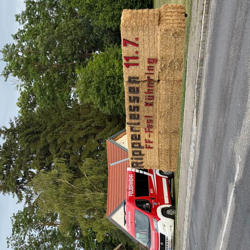 A fire truck is parked on the side of a road, with a hay bale featuring the word 'Kupferlesen' behind it. Trees and a building are visible in the background.