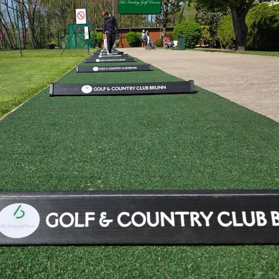 Ein Golfspieler übt auf einem Golfplatz mit Kunstrasen, mit mehreren Golf-Übungszielen, die mit 'Golf & Country Club Brunn' beschriftet sind. Im Hintergrund befinden sich Bäume und ein Gebäude.