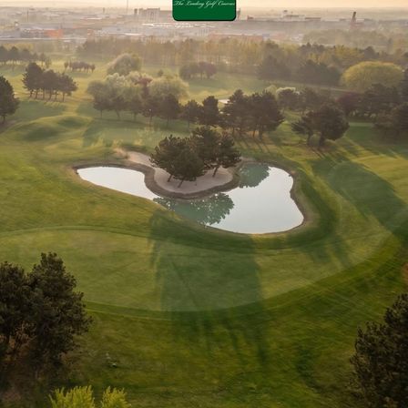 Luftaufnahme eines Golfplatzes mit einem herzförmigen Teich und Bäumen. Der Himmel ist klar mit einem Hauch von Nebel.