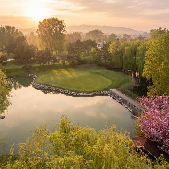 Ein ruhiger Golfplatz mit einem Teich, umgeben von üppigem Baumbestand und einem malerischen Sonnenuntergang im Hintergrund.