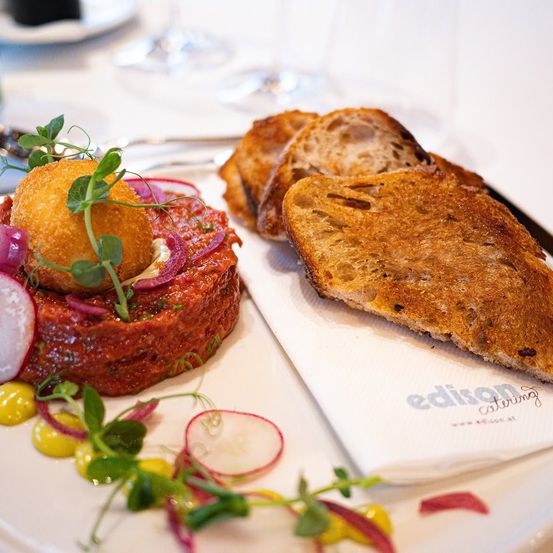 Ein Teller mit einem roten Gericht, gekrönt von einer frittierten Kugel, garniert mit grünen Blättern und kleinen roten Zwiebeln, begleitet von getoastetem Brot und einer Serviette mit dem Text 'edison catering'.