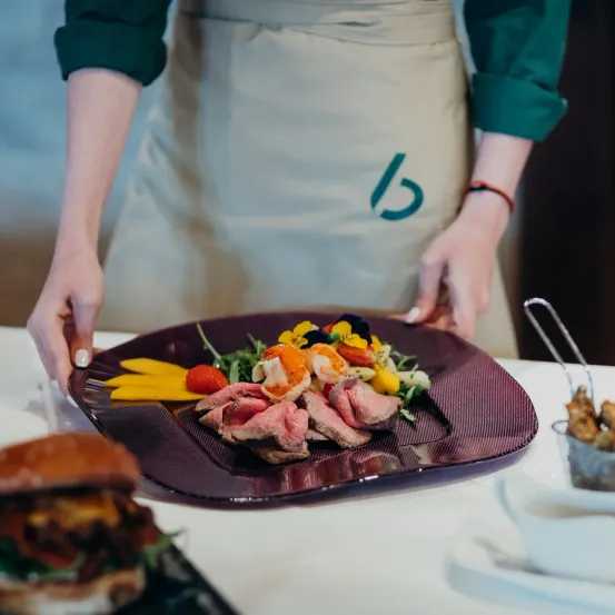 Ein Koch hält einen Teller mit einem Gericht aus Fleisch, Garnelen und Gemüse auf einem weißen Tisch.