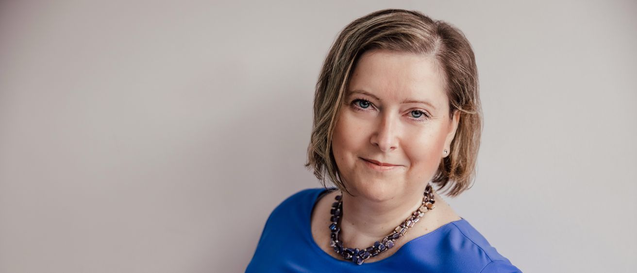 Eine Frau mit kurzen braunen Haaren, die ein blaues Oberteil und eine Perlenkette trägt, posiert für ein Porträt vor weißem Hintergrund.