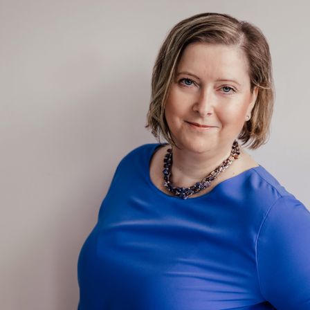 Eine Frau mit kurzen Haaren lächelt und trägt ein blaues Oberteil und eine Halskette. Sie steht vor einer weißen Wand.