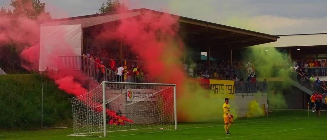 Ein Fußballspieler steht neben dem Tor, mit rotem Rauch und einer Menge dahinter. Es gibt ein Gebäude mit einem Banner und gelbem Rauch, der von der Seite kommt.
