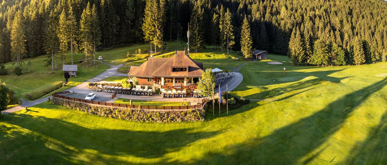 Eine Luftaufnahme eines Golfplatzes mit einem Clubhaus, saftigem grünem Gras und Bäumen im Hintergrund. Flaggen säumen den Weg zum Clubhaus.