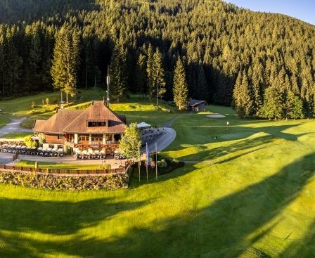 Ein Luftbild eines Golfclubs mit einem großen Gebäude, umgeben von einem üppigen grünen Feld und dichtem Wald mit einem Berg im Hintergrund.