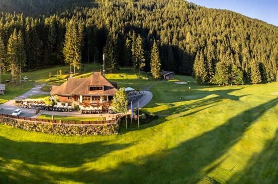 Ein Luftbild eines Golfclubs mit einem großen Gebäude, umgeben von einem üppigen grünen Feld und dichtem Wald mit einem Berg im Hintergrund.