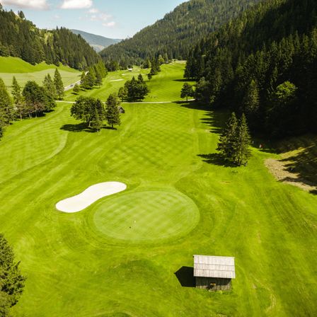 Luftaufnahme eines Golfplatzes mit saftig grünem Gras, Bäumen und einem kleinen Holzhaus in der Ferne. Berge und ein bewölkter Himmel im Hintergrund.