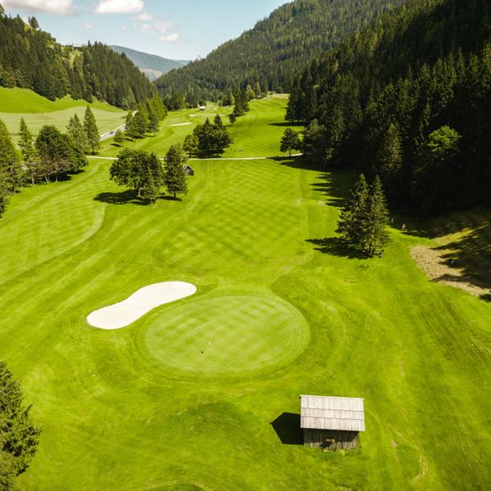 Luftaufnahme eines Golfplatzes mit saftig grünem Gras, Bäumen und einem kleinen Holzhaus in der Ferne. Berge und ein bewölkter Himmel im Hintergrund.