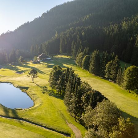 Ein friedlicher Golfplatz in den Bergen mit einem kleinen Teich, umgeben von üppigem Grün und Bäumen, unter einem klaren blauen Himmel.