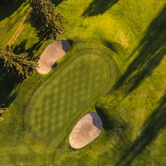 Eine Luftaufnahme eines Golfplatzes mit zwei Sandbunkern und saftigem grünem Gras, das Schatten auf den Boden wirft.