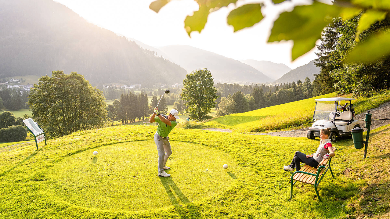 Ein Golfer in einem grünen Shirt schwingt seinen Schläger auf einem Golfplatz mit Bergen im Hintergrund.