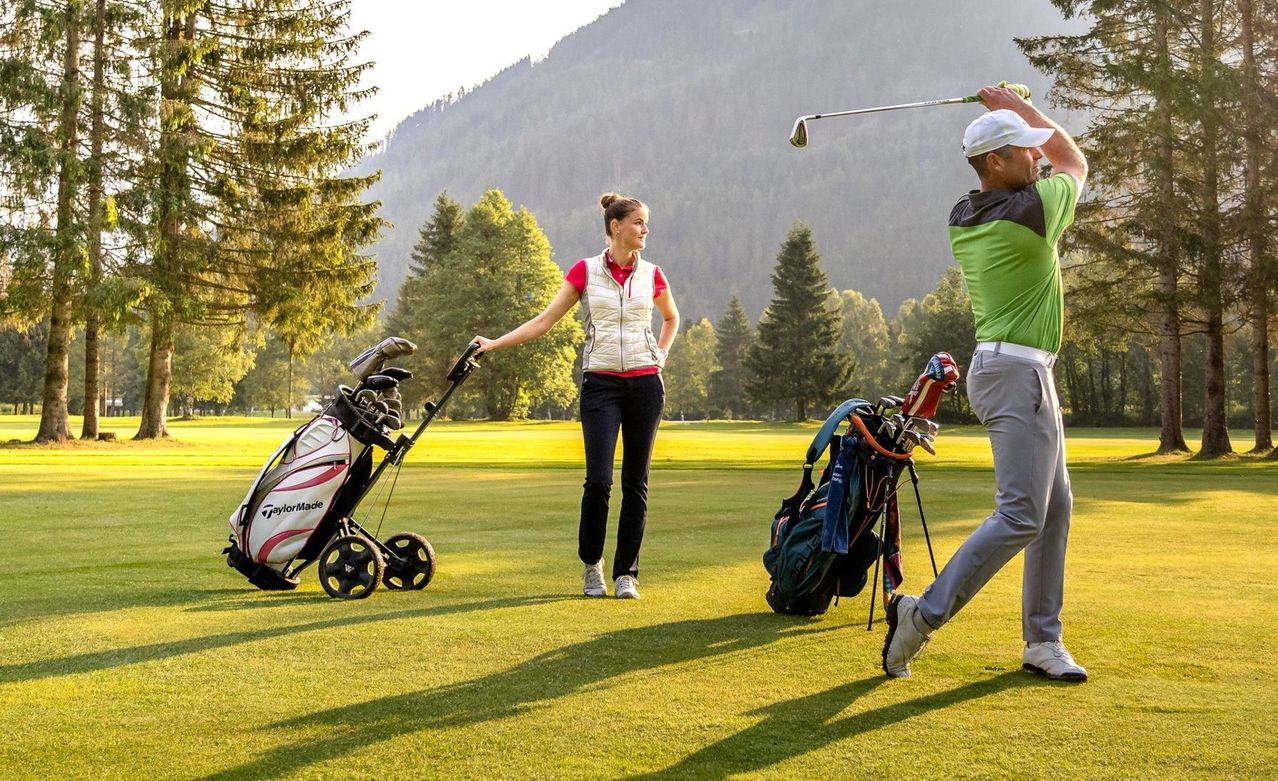 A man and woman play golf on a sunny day. The man swings his club, while the woman holds a club and a golf bag.