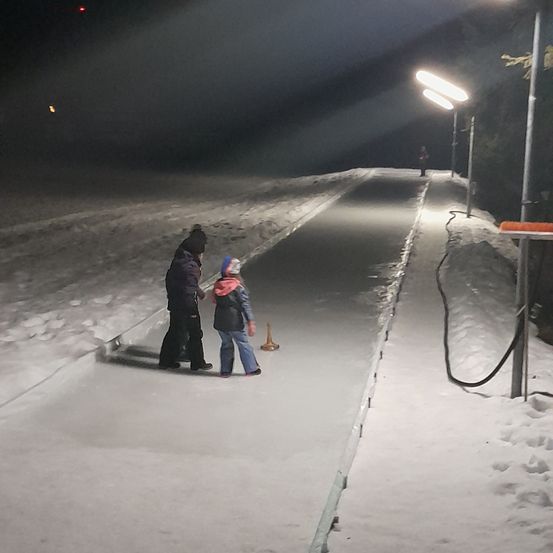 Zwei Personen stehen auf einer beleuchteten Eislaufbahn, eine hält einen Kegel. Schnee bedeckt die Seiten, und Straßenlaternen säumen den Weg.