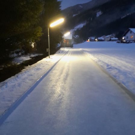 Eine verschneite Straße mit Straßenlaternen und einem Berg im Hintergrund bei Nacht.
