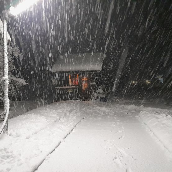 Eine kleine Hütte wird inmitten starken Schneefalls bei Nacht von warmen Lichtern beleuchtet. Schneebedeckte Bäume und eine Straßenlaterne verstärken die Winterszene.