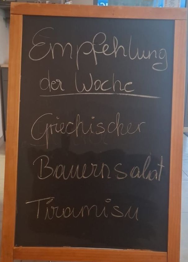 Ein Tafelmenü-Board zeigt 'Empfehlung der Woche' mit 'Griechischer Bauernsalat' und 'Tiramisu' als wöchentliche Empfehlungen.