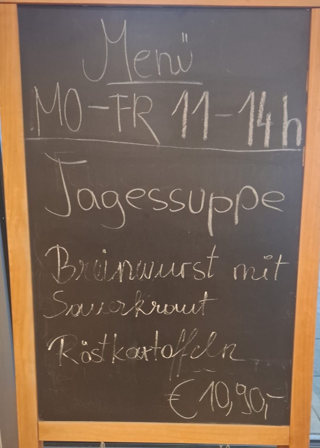 Eine Kreidetafel mit weißer Kreideschrift. Oben steht 'Geöffnet', darunter 'Mo-Fr 11-14 Uhr', in der Mitte 'Tagessuppe' und darunter 'Brühwurst mit Sauerkraut und Röstkartoffeln'.
