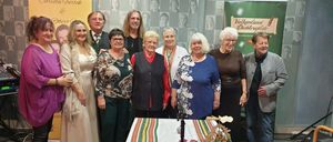 Eine Gruppe von Menschen, darunter mehrere Frauen, steht hinter einem Tisch mit einem Banner, auf dem 'Vulkanland Dichtergilde' steht. Die Frau in der Mitte trägt eine Halskette und eine Uhr. Einige Leute tragen Brillen und Medaillen.