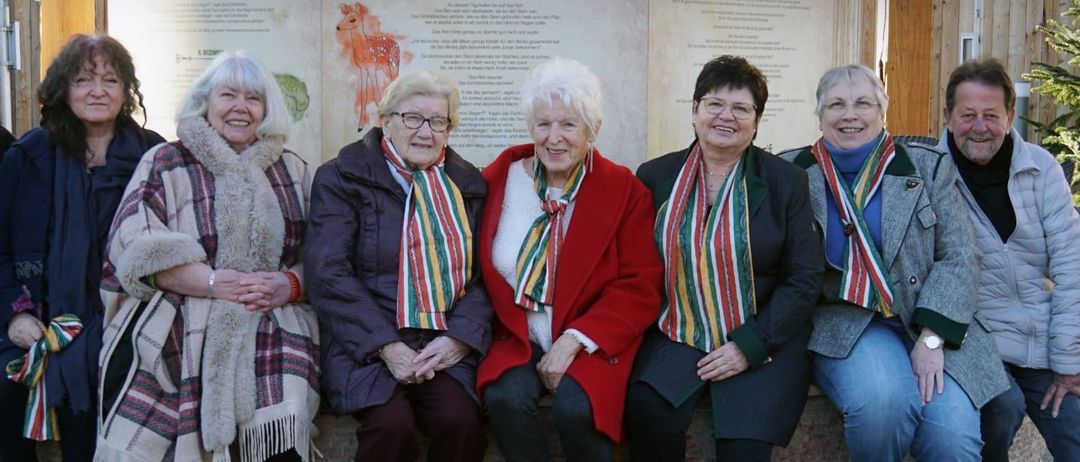 Eine Gruppe von Frauen in Winterkleidung posiert für ein Foto vor einer Wand mit Schrift und Illustrationen. Sie tragen bunte Schals.