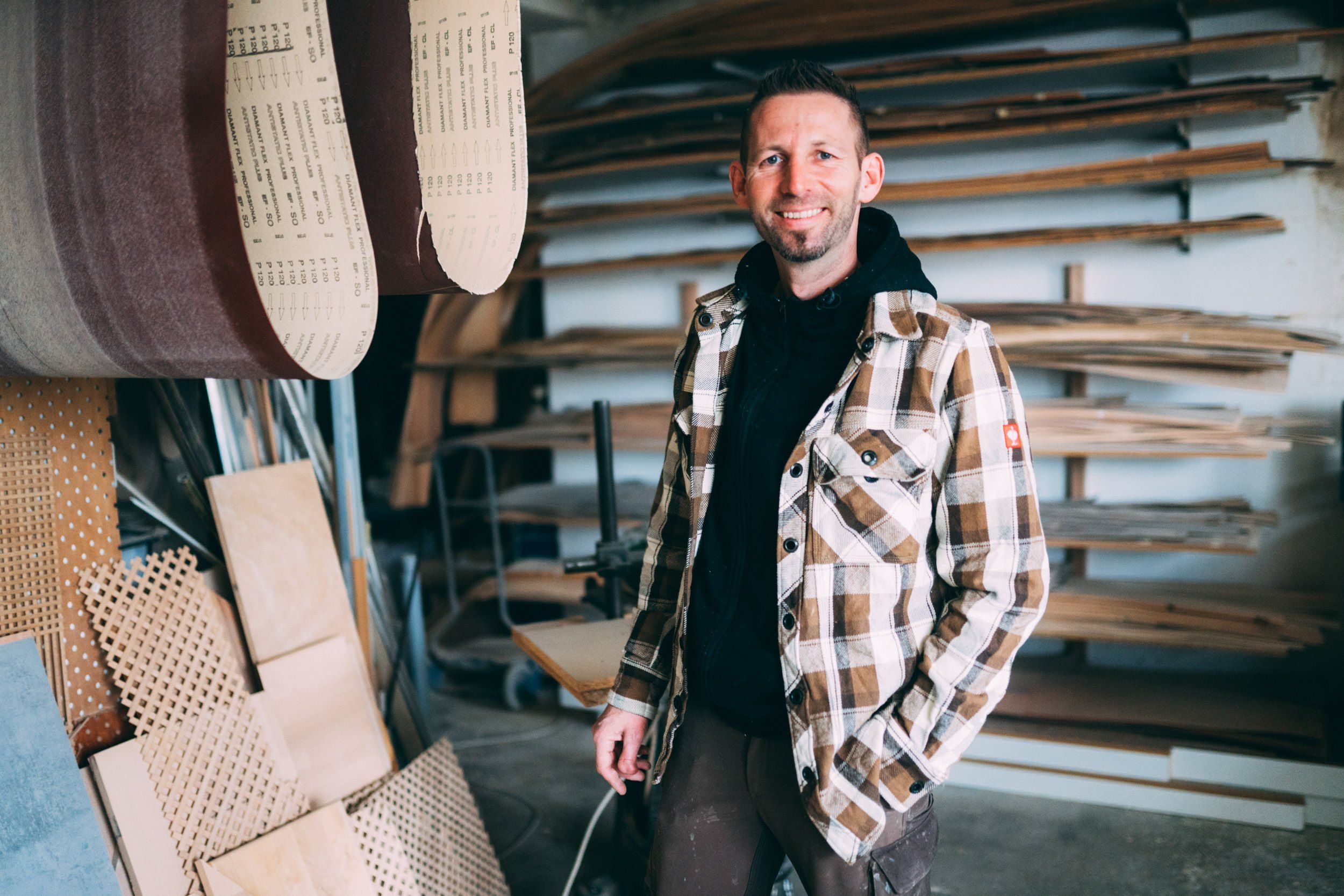 Ein Mann steht lächelnd in einer Werkstatt und trägt eine karierte Jacke. Hinter ihm sind Regale mit Holzbrettern und Werkzeugen zu sehen, und Materialien hängen von der Decke herab.