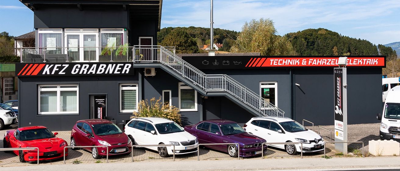 Ein Werkstattgebäude mit dem Schriftzug Technik & Fahrner, mehrere Autos davor geparkt, eines rot, zwei weiß und eines lila.