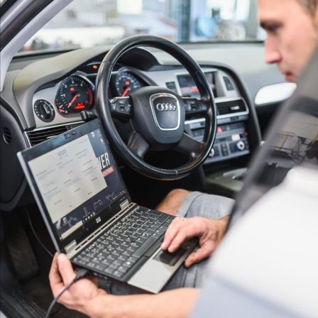 Ein Mann sitzt in einem Audi-Auto und benutzt einen Laptop, der mit dem Fahrzeugsystem verbunden ist und Text auf dem Bildschirm anzeigt.