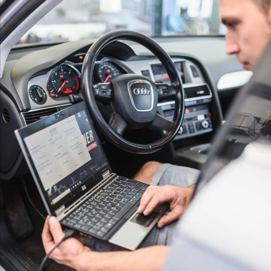 Ein Mann sitzt in einem Audi-Auto und benutzt einen Laptop, der mit dem Fahrzeugsystem verbunden ist und Text auf dem Bildschirm anzeigt.