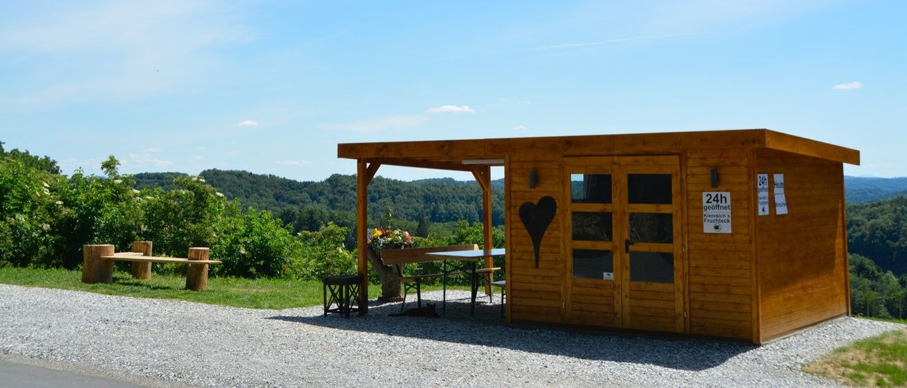 Ein Holzschuppen mit einem schwarzen Herz auf der Tür steht an einem malerischen Ort mit einem Tisch, Stühlen und einer Bank draußen unter einem Dach. In der Nähe gibt es eine Bank und einen Baumstumpf auf dem Rasen. Der Hintergrund zeigt einen klaren blauen Himmel mit einigen Wolken und einem entfernten Bergrücken.