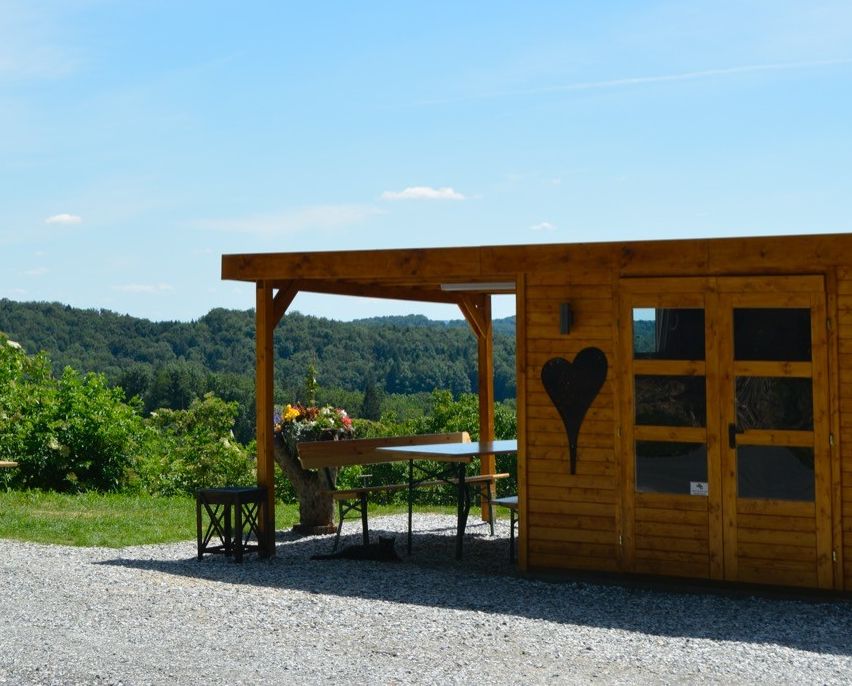 Ein Holzschuppen mit einem schwarzen Herz auf der Tür steht an einem malerischen Ort mit einem Tisch, Stühlen und einer Bank draußen unter einem Dach. In der Nähe gibt es eine Bank und einen Baumstumpf auf dem Rasen. Der Hintergrund zeigt einen klaren blauen Himmel mit einigen Wolken und einem entfernten Bergrücken.