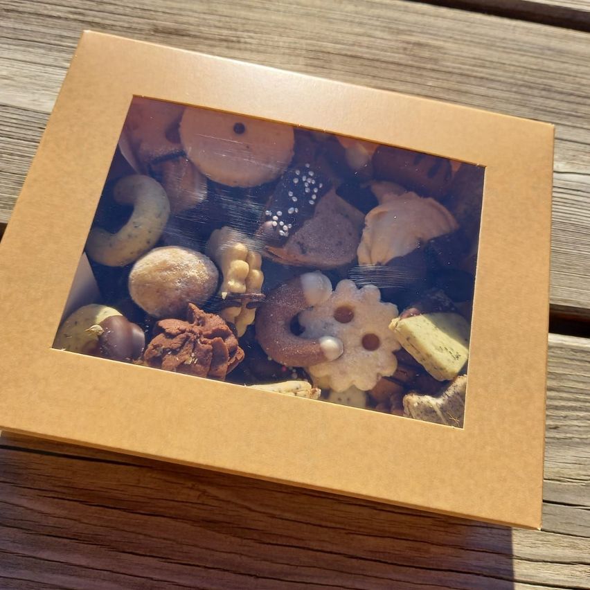 Eine braune Box mit einem Glasfenster zeigt eine Auswahl an Keksen und Gebäck. Das Sortiment umfasst Schokoladenüberzogene Kekse, zuckerbestreute Donuts und kunstvoll gestaltete Kekse.