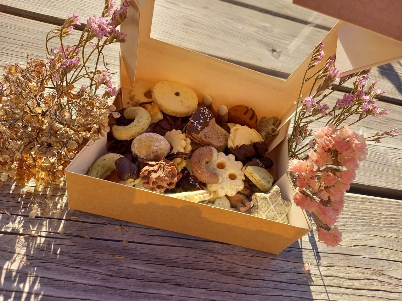 Eine Schachtel mit verschiedenen Keksen und Gebäck, darunter Schokoladenüberzogene, mit Streuseln und blumenförmige Kekse, auf einem Holztisch mit Blumen.