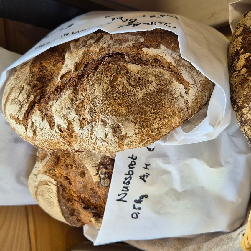 Zwei Brotlaibe sind in weißes Papier gewickelt und auf einer Holzoberfläche platziert. Ein Laib ist mit 'Nußbrot' beschriftet, während der andere einen handgeschriebenen Aufkleber hat.