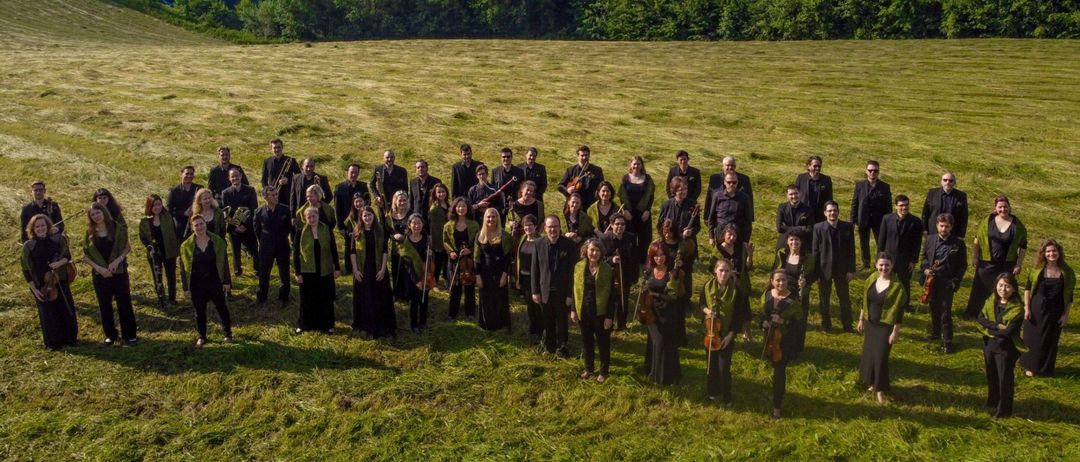 Eine Gruppe von Musikern und Sängern steht auf einer Wiese mit Bergen und einer Stadt im Hintergrund.