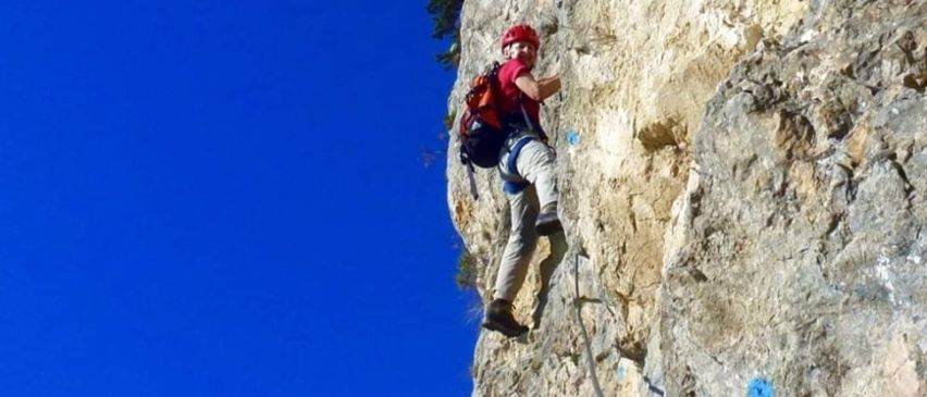 Eine Person mit rotem Helm und Rucksack klettert an einer steilen Felswand mit einem Seil nach oben.