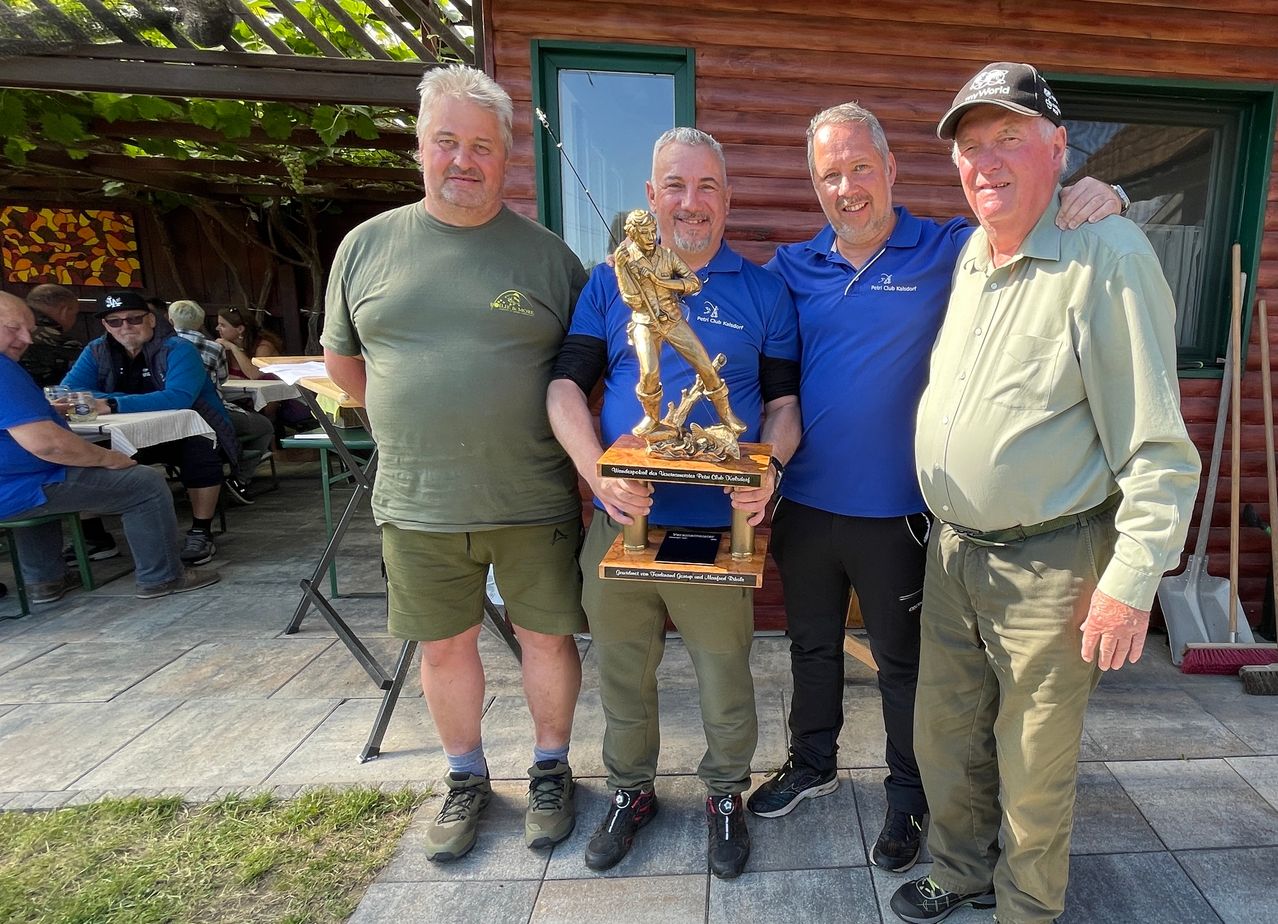 Vier Männer posieren mit einer Trophäe draußen, zwei tragen blaue Hemden und einer hält eine Baseballtrophäe in den Händen.
