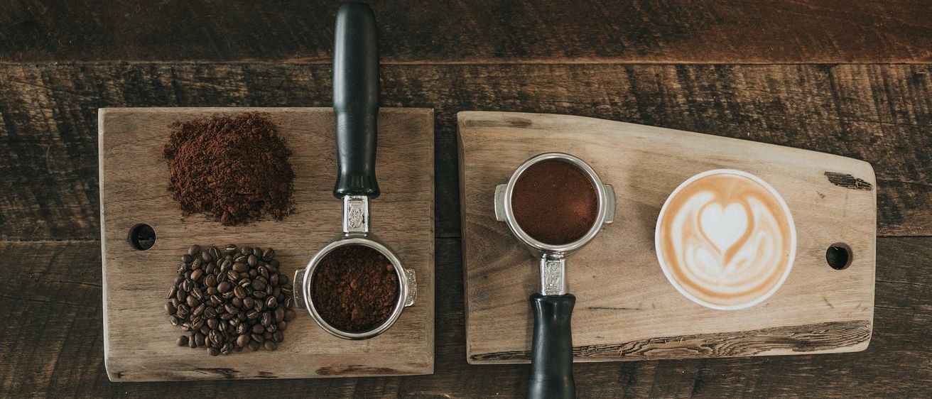 Ein Holztisch zeigt Kaffeebohnen, eine Kaffeepresse und einen Latte mit Herzdesign.