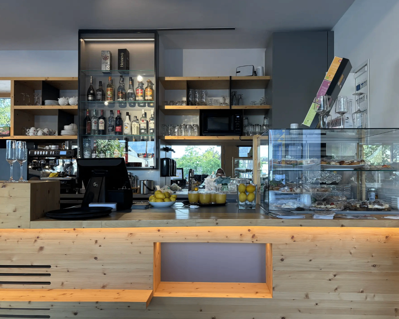 Ein modernes Café mit einer Holztheke, einer Vitrine voller Gebäck und Regalen, die mit Flaschen und Gläsern bestückt sind.