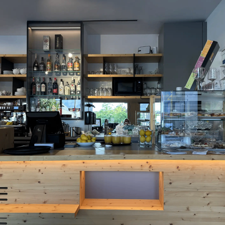 Ein modernes Café mit einer Holztheke, einer Vitrine voller Gebäck und Regalen, die mit Flaschen und Gläsern bestückt sind.
