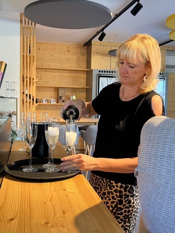 A woman is pouring champagne into glasses on a wooden table in a modern kitchen.