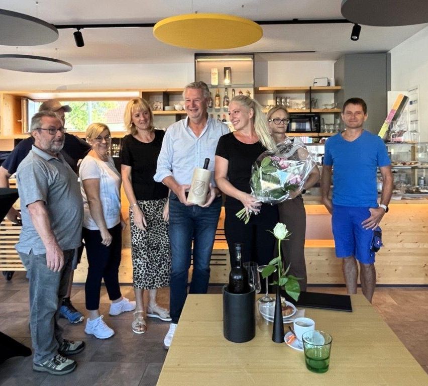 A group of people stand together in a room, some holding flowers and bottles. The man in the center holds a bottle, and a woman next to him holds a bouquet of flowers. There is a table with wine bottles and a vase with a flower in the foreground.