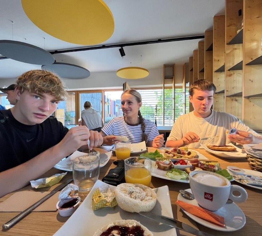 Three people are sitting at a table in a restaurant, enjoying their breakfast. The table is filled with various foods, including bread, fruit, and coffee. The atmosphere is bright and cheerful.